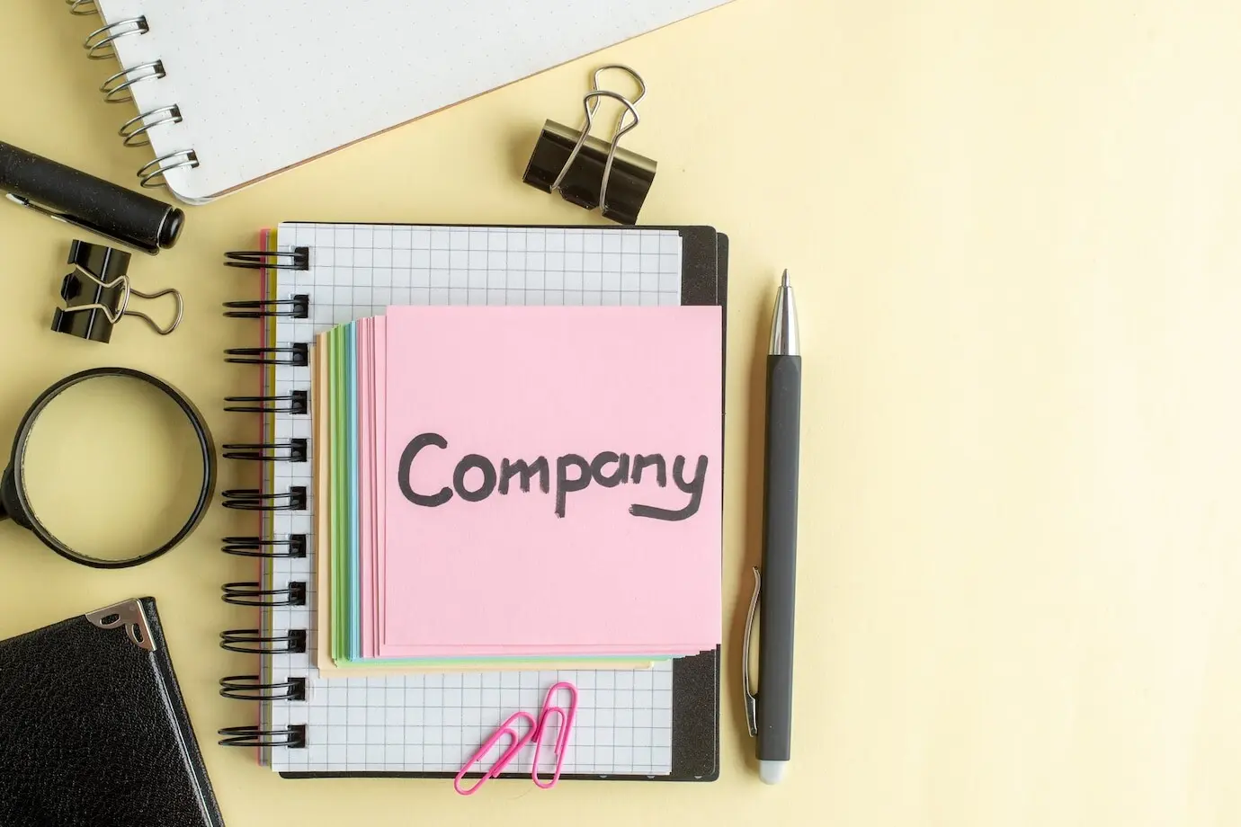 Вид сверху на светлую поверхность: записка с надписью «company», маленькие разноцветные бумажные стикеры, блокнот, тетрадь, ручка, работа, труд, школа, офис, бизнес, деньги, цвет.