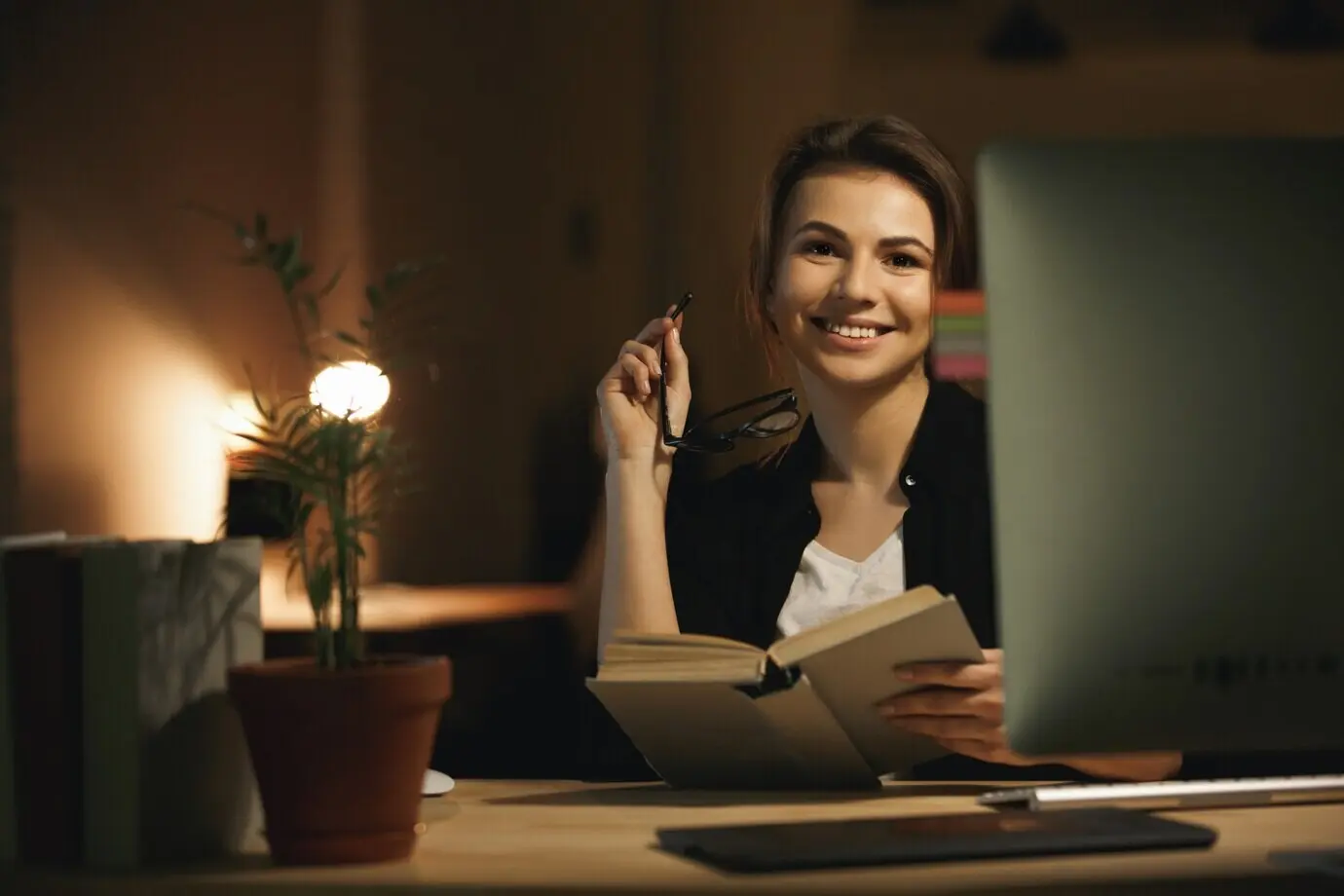 Счастливая молодая женщина-дизайнер пользуется компьютером и держит книгу.